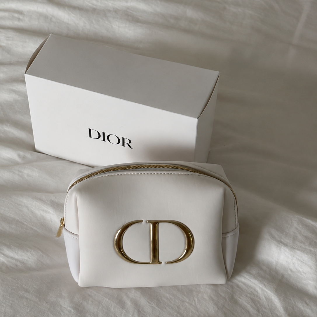 A white Dior cosmetic pouch with a gold-tone CD logo, placed in front of its white Dior box on a white fabric surface.