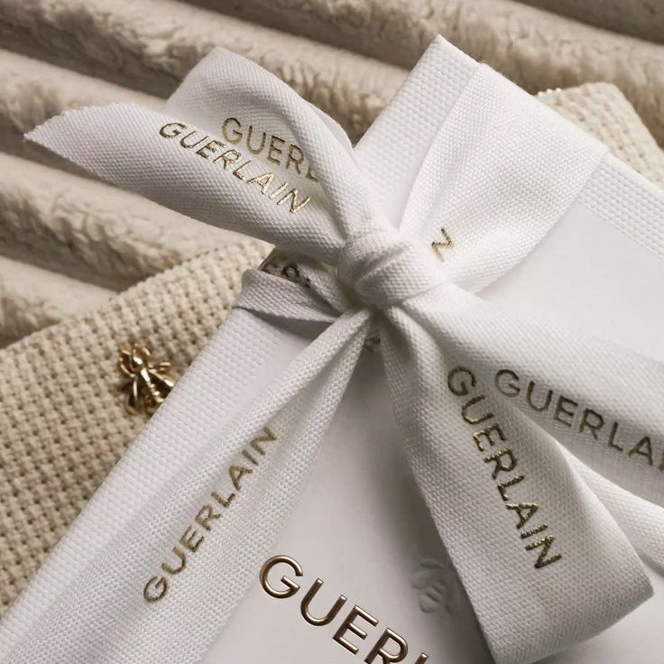A close-up of a white Guerlain Paris gift box tied with a white ribbon printed with the brand name, resting on a textured beige fabric background.