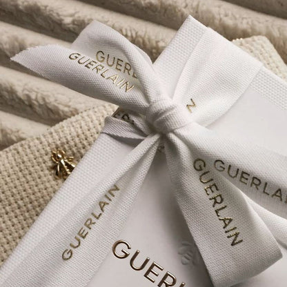 A close-up of a white Guerlain Paris gift box tied with a white ribbon printed with the brand name, resting on a textured beige fabric background.