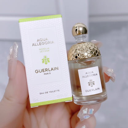 A hand holds a bottle of Guerlain Aqua Allegoria Nerolia Vetiver Eau de Toilette next to its white packaging box with light green accents. The bottle features a textured gold cap and a clear glass body showing the pale amber liquid inside.