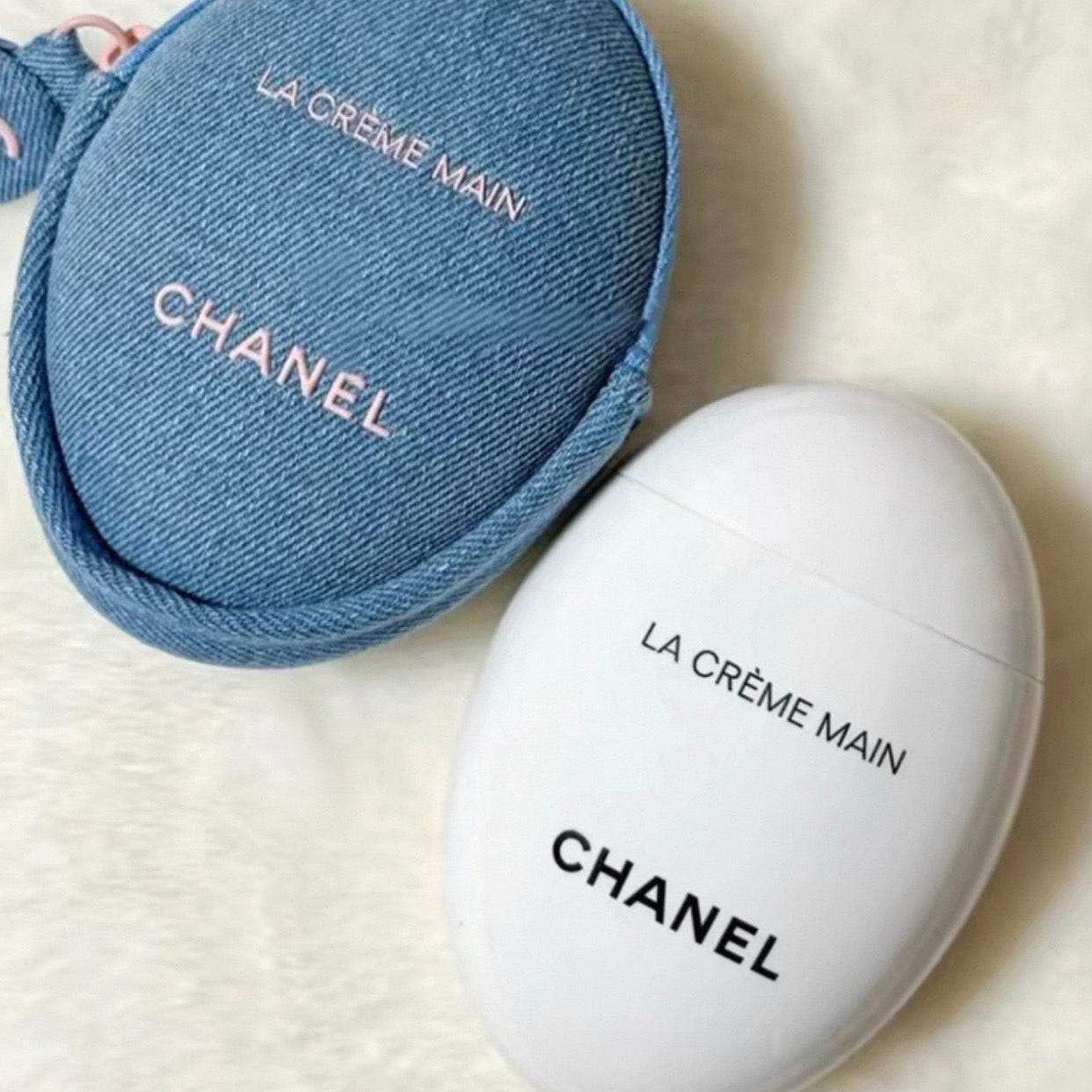A white oval Chanel La Crème Main cosmetic container next to a blue denim zippered pouch with pink lettering, on a soft white textured background.