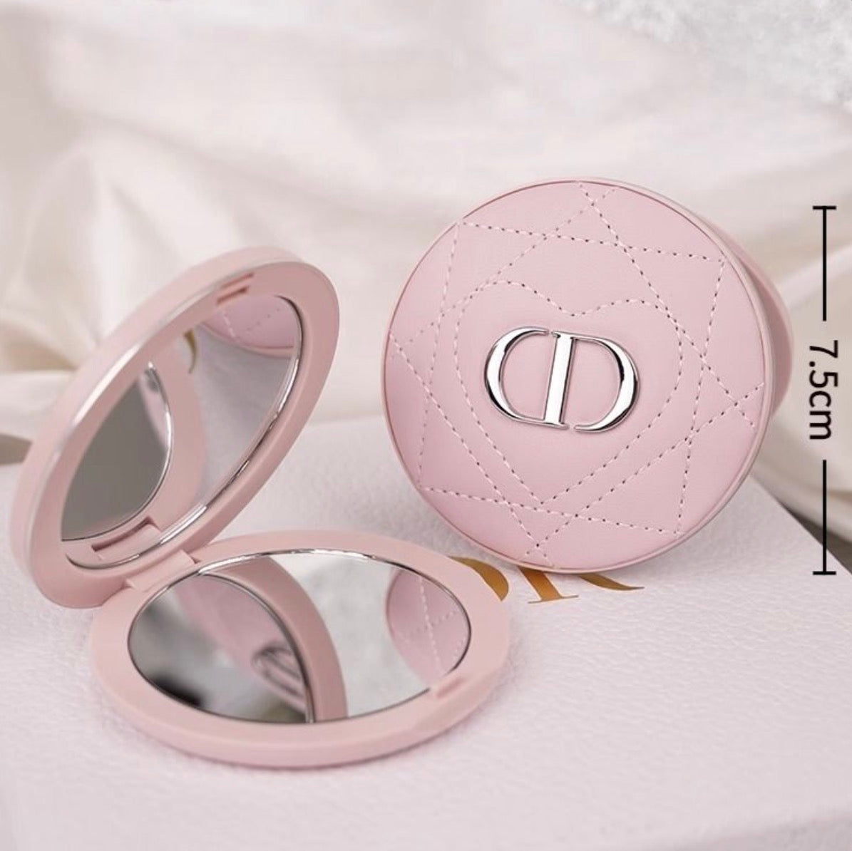 A pink round compact shown open and closed on a soft white surface. The closed compact displays a quilted pattern and a silver-tone interlocking D logo, while the open compact reveals mirrored interiors.