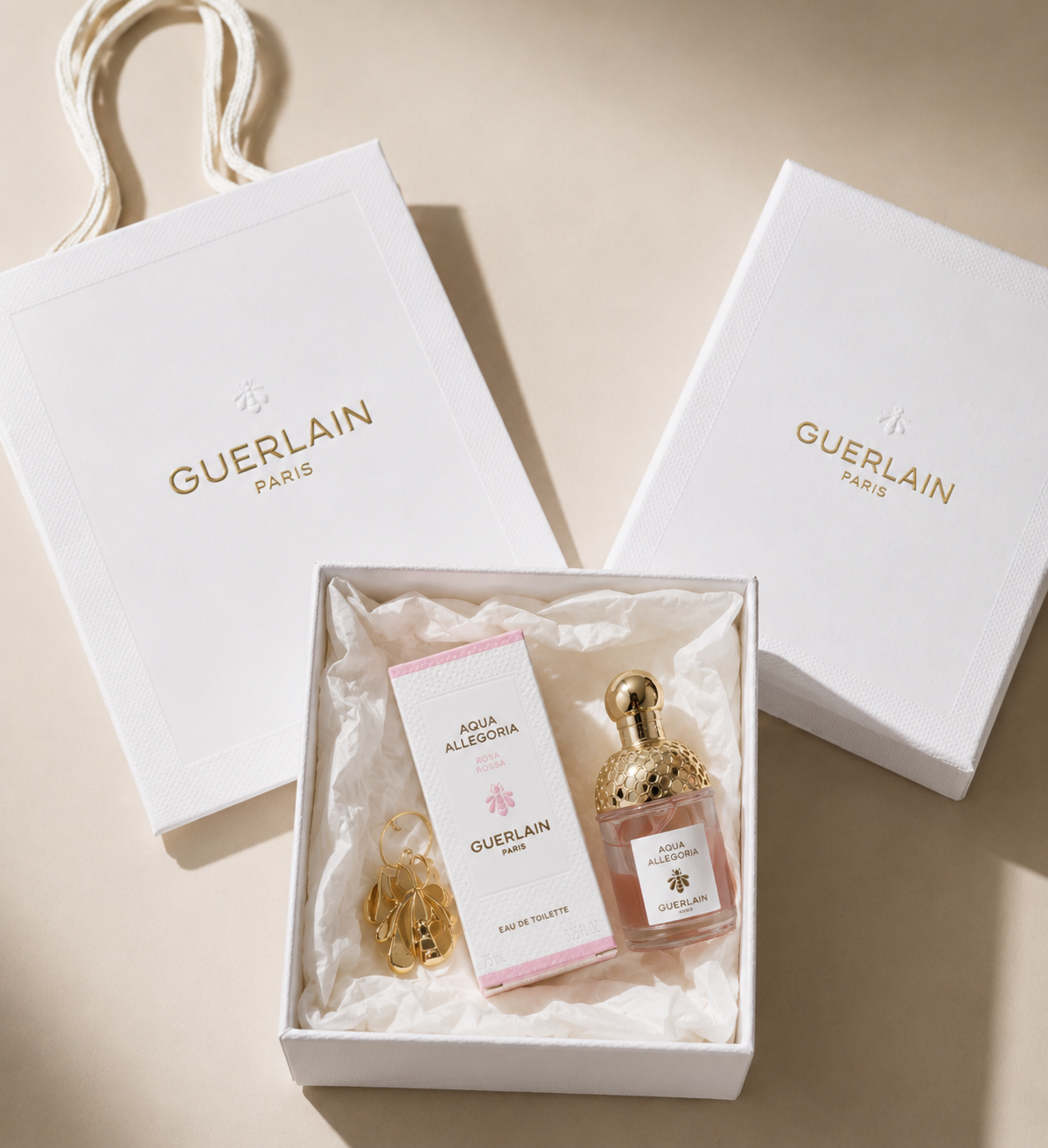 Guerlain Agressif Orchide perfume bottle with a gold cap, displayed in a white gift box with a pink card and gold earrings, alongside white Guerlain shopping bags and a box lid on a beige background.