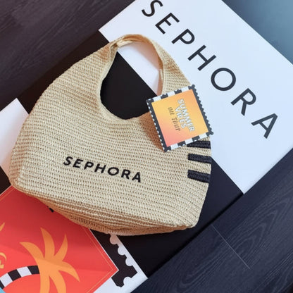 A woven straw tote bag in beige with black SEPHORA branding, placed on top of a large white SEPHORA box. The bag has black-and-white striped side accents and a hangtag that reads 'SEPHORA Summer Sale 2025'.