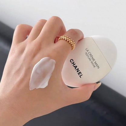 A hand holding a white Chanel La Crème Min bottle next to a smear of white cream, with a gold ring on one finger.