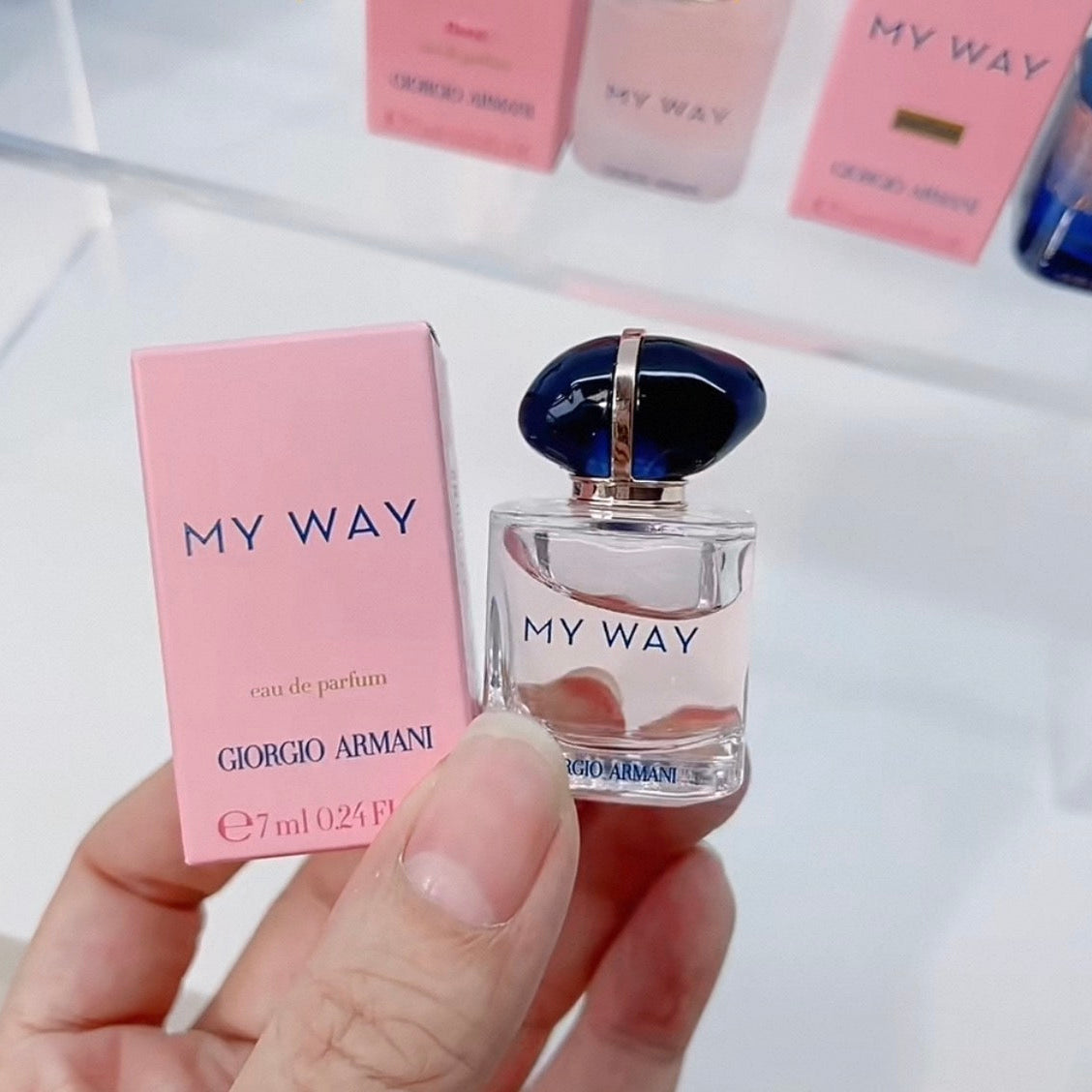 A hand holds a small Giorgio Armani 'My Way' perfume bottle next to its pink box, with more matching pink boxes blurred in the background.