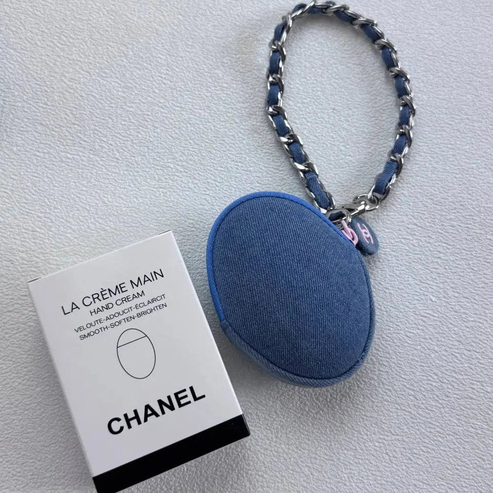 A Chanel La Crème Main hand cream bottle with its original white and black packaging box, displayed on a light gray textured surface. The bottle features a blue fabric-covered oval body with a silver chain strap.