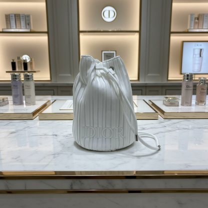 A white pleated drawstring bucket bag with an embossed Dior logo on the front, resting on a beige fabric surface.