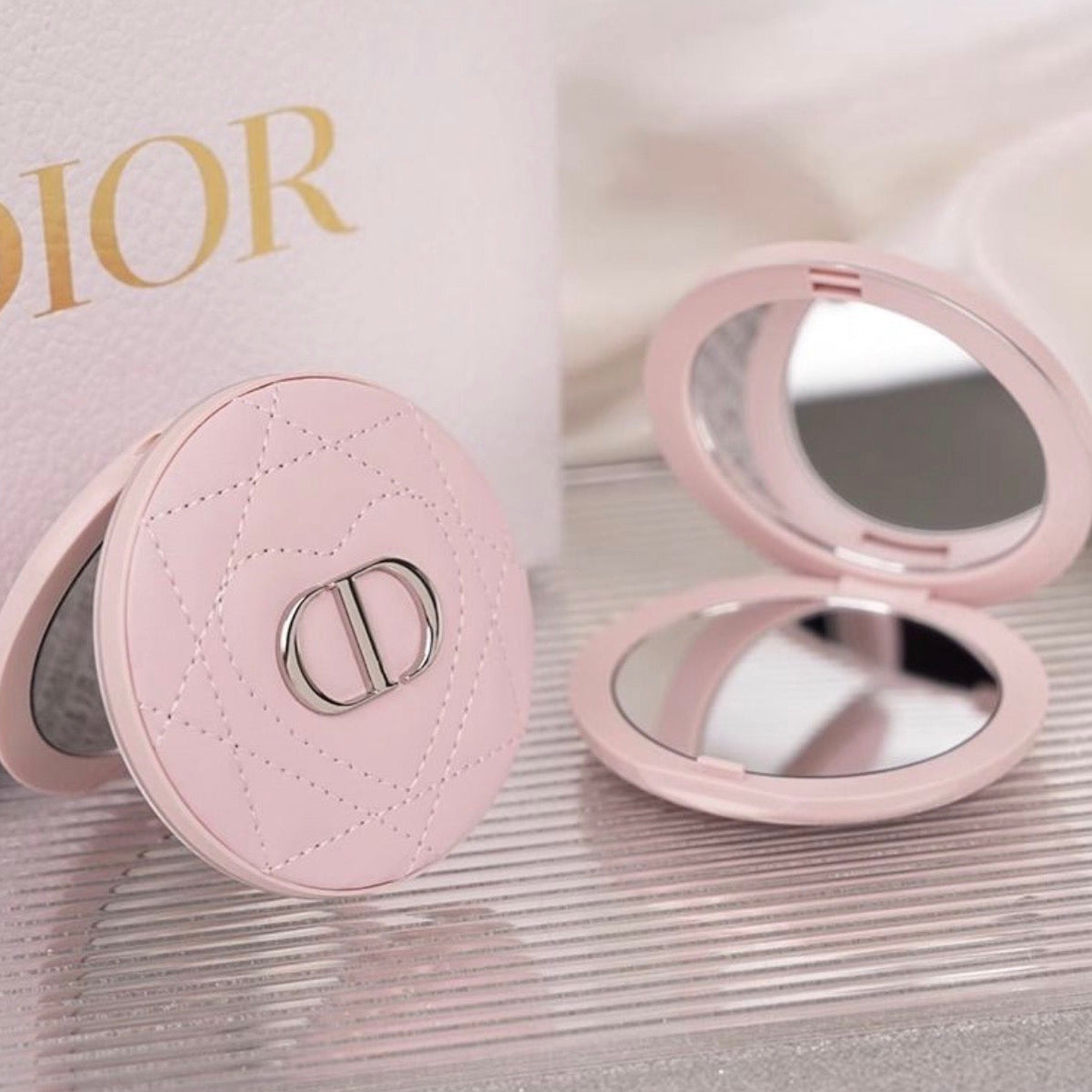 A pink Dior compact mirror shown closed and open on a reflective surface, with a Dior-branded box partially visible in the background.