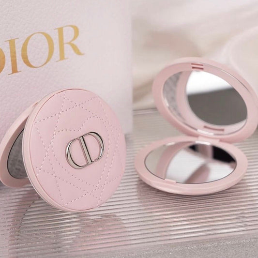 A pink Dior compact mirror shown closed and open on a reflective surface, with a Dior-branded box partially visible in the background.