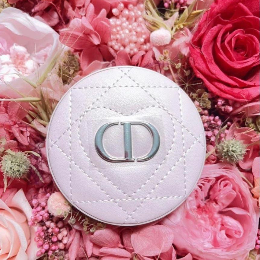 A round white quilted cosmetic compact with a silver-tone interlocking CD logo, placed on a dense bed of pink and red roses.