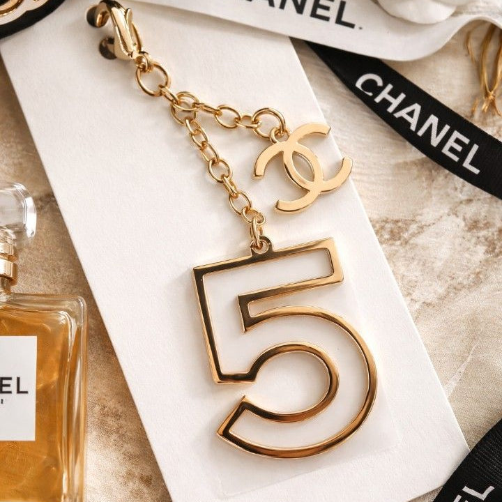 Gold number 5 keychain with Chanel branding on a marble surface with Chanel perfume bottle and white flower.