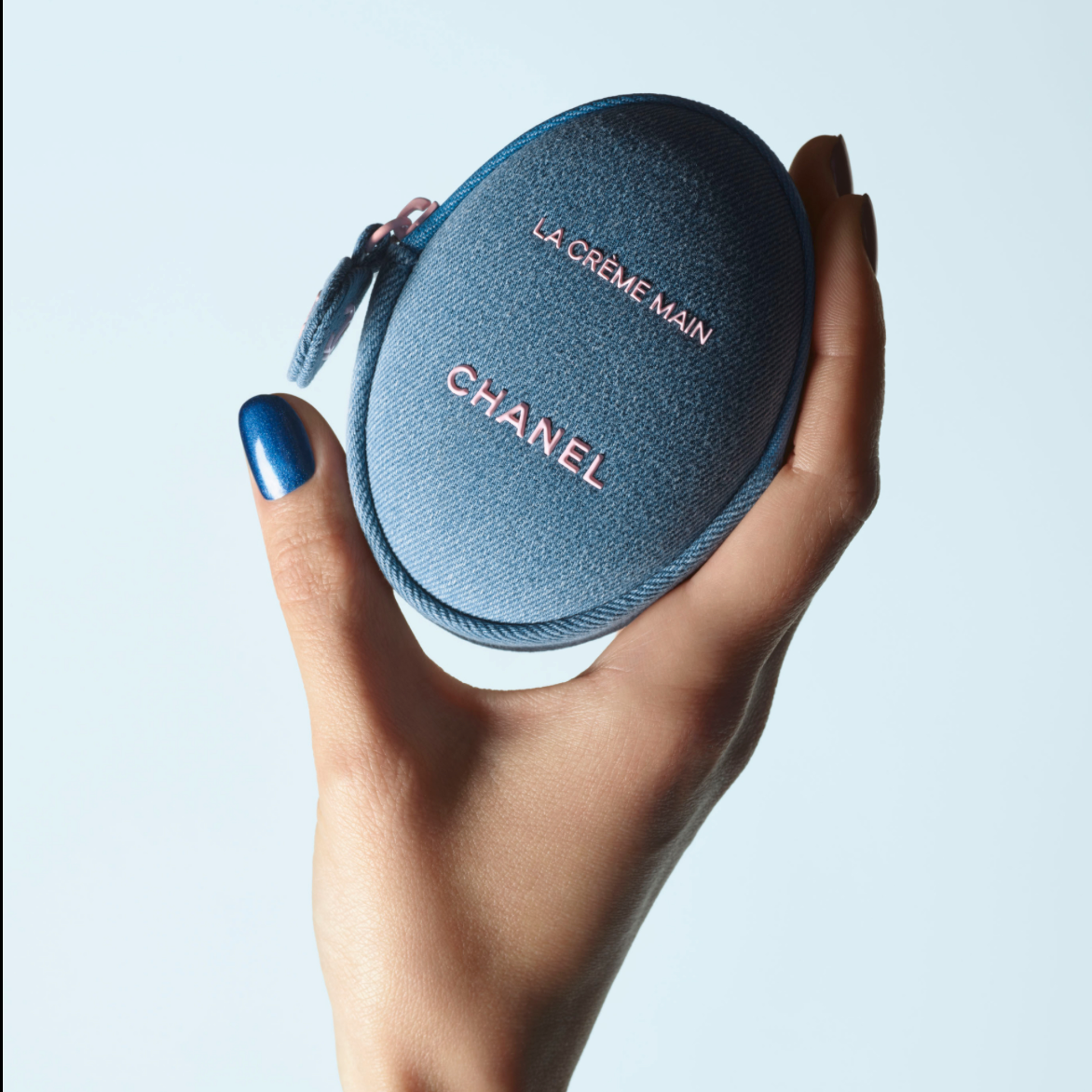 A hand with blue nail polish holds a round blue Chanel cosmetic case with the words 'LA CREME MAIN' and 'CHANEL' in pink lettering, against a light gray background.