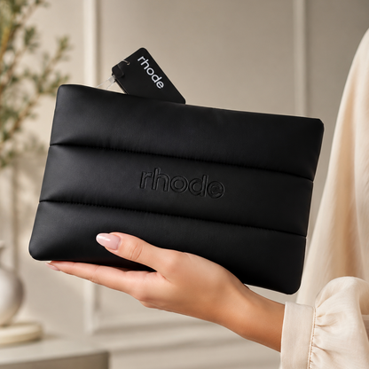 A person in a cream-colored blouse holds a black padded zip pouch with the rhode logo embossed on the front. The pouch has horizontal quilted stitching and a black hanging tag with the rhode brand name.