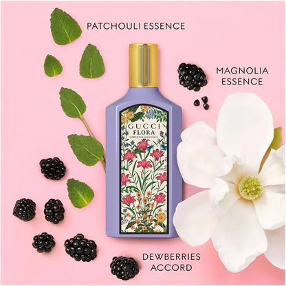 A Gucci Flora patchouli essence bottle with a lavender body, gold cap, and floral label, arranged on a pink background with green leaves, blackberries, and a white magnolia flower. Text labels indicate patchouli essence, magnolia essence, and dewberries accord.
